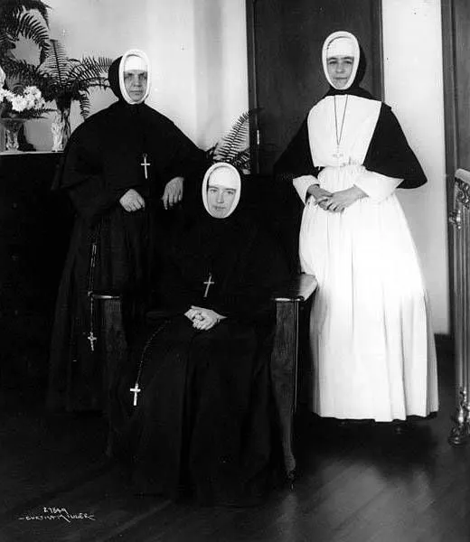 Tres monjas con hábitos largos, la cabeza cubierta por el velo, dos de pie y una sentada