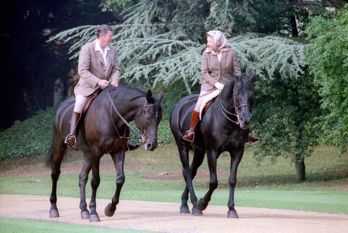 La reina Isabel II a caballo, con un pañuelo en la cabeza atado bajo la barbilla, junto al presidente Reagan